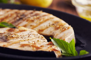Grilled swordfish steaks, cooked justabout perfectly.lices in a cast iron pan on a wooden table, garnished with mint, oregano, salt and salmoriglio